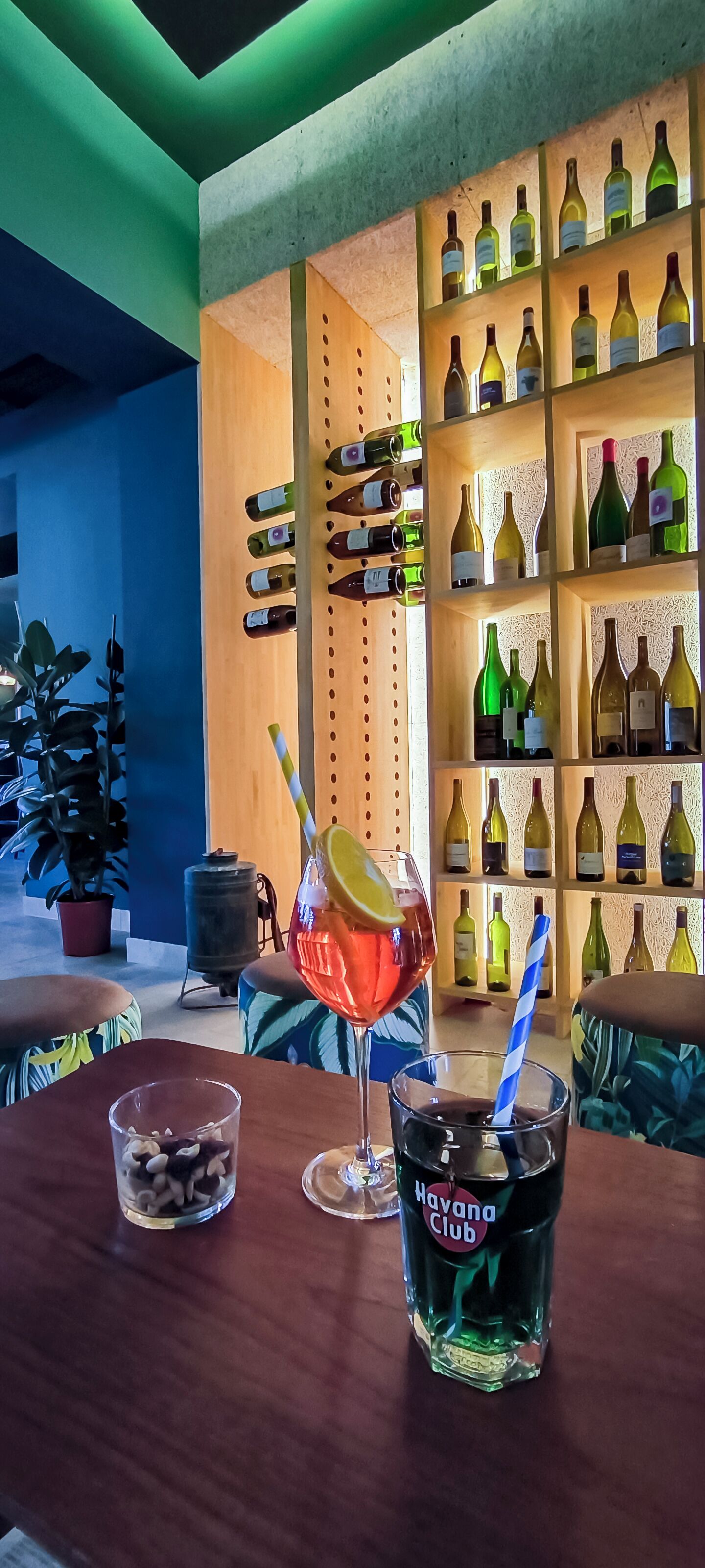 A bar area with a wooden shelf displaying various wine bottles, a table with drinks, and a potted plant.