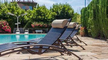 A row of sun loungers with cushions by a pool, a hotel sign in the background, and lush greenery.