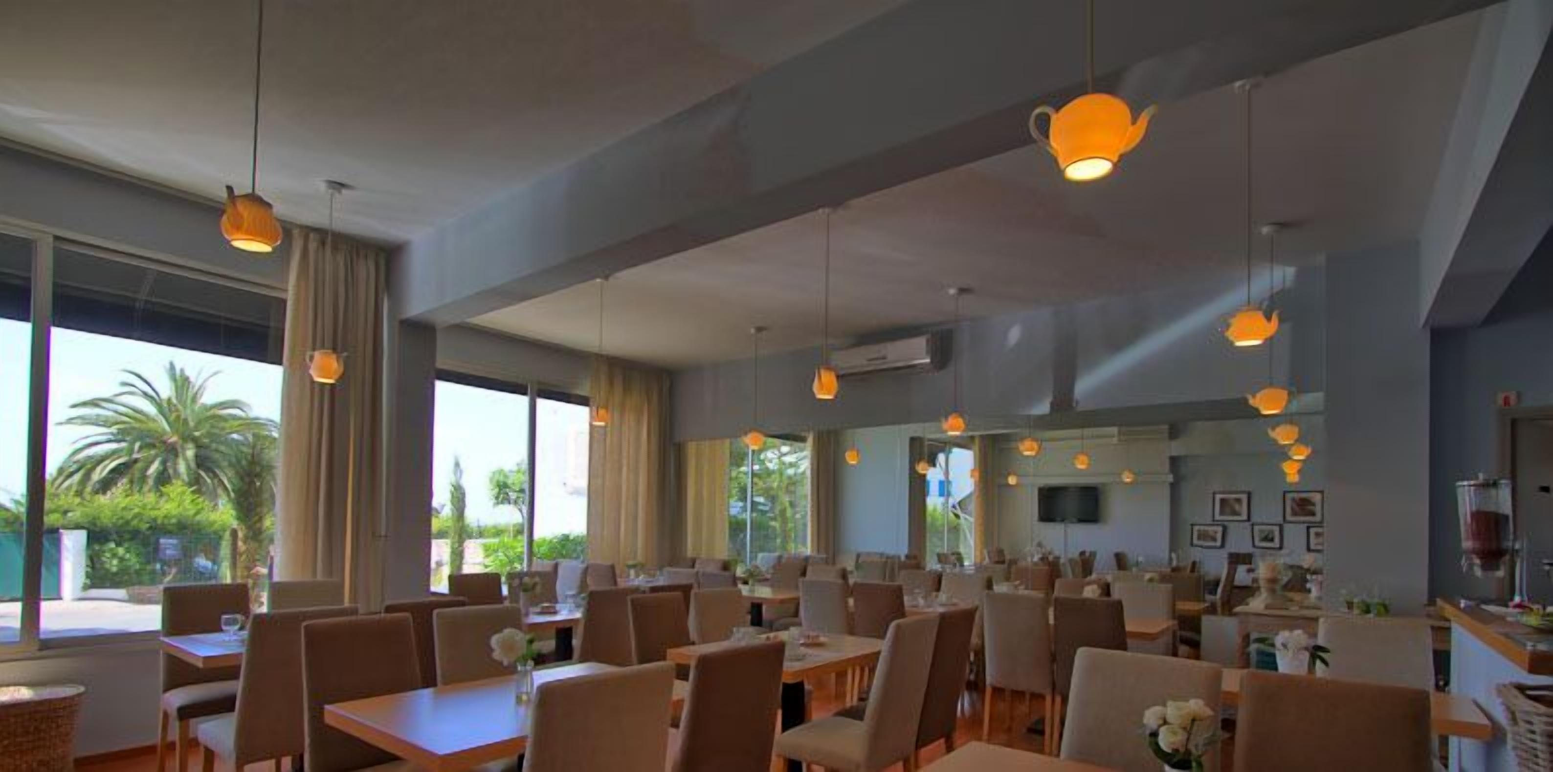 A spacious restaurant with multiple dining tables and chairs, large windows offering views of greenery, and pendant lighting.