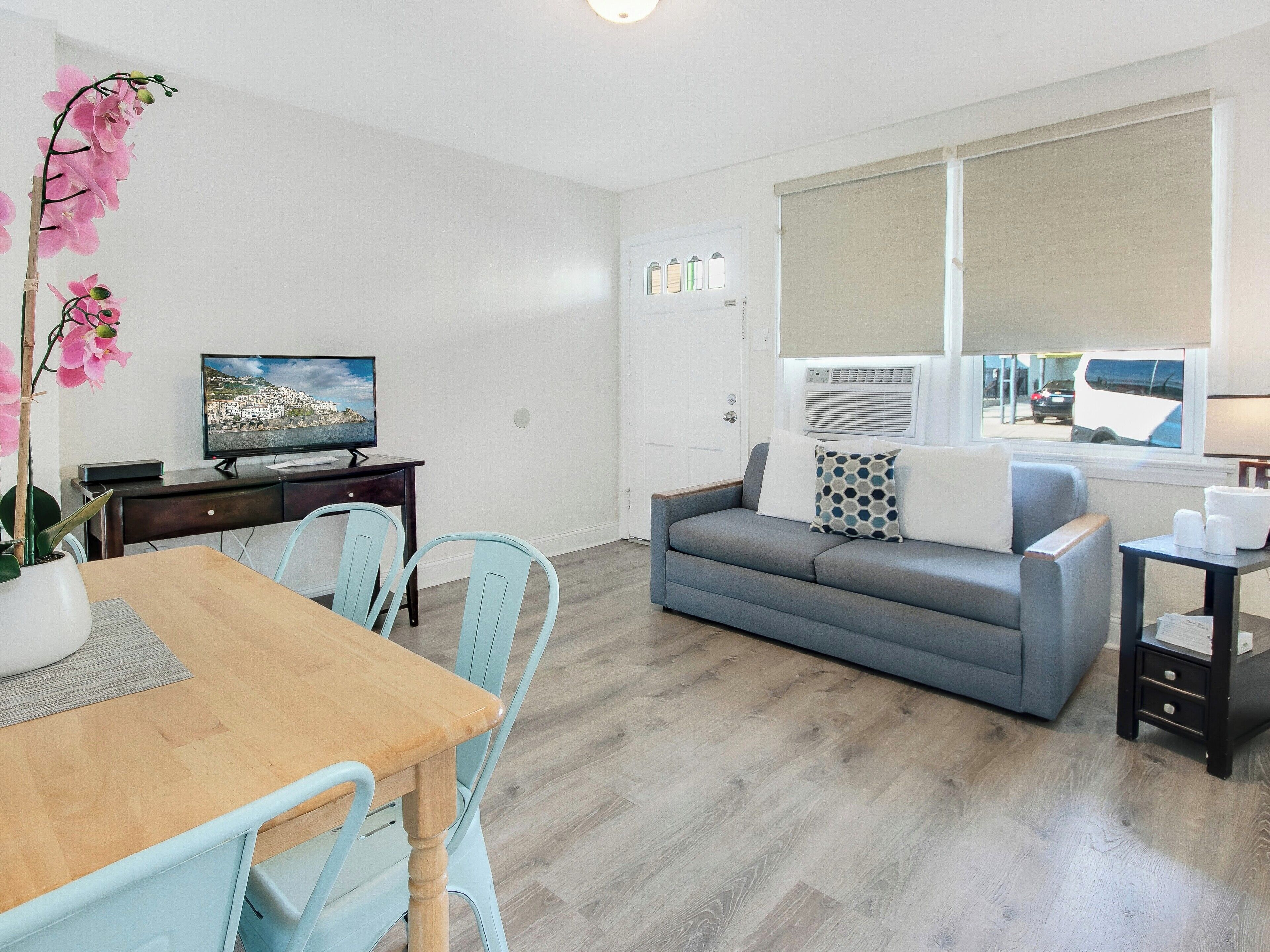 A modern living room with a grey sofa, a wooden dining table with chairs, a TV, and a large window with blinds.