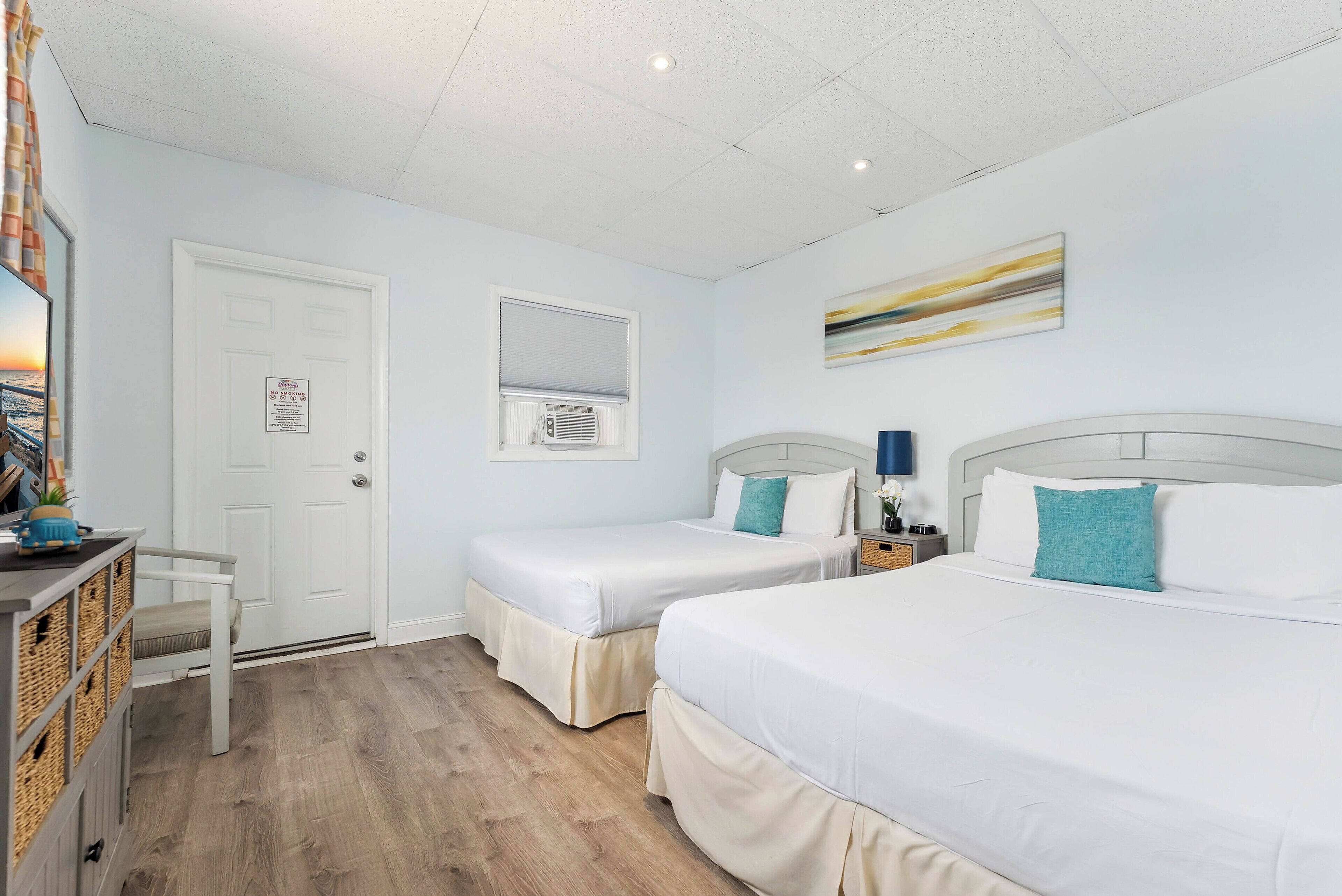 Une chambre d’hôtel avec deux lits, un bureau, une chaise, une fenêtre avec stores et un climatiseur fixé au mur.