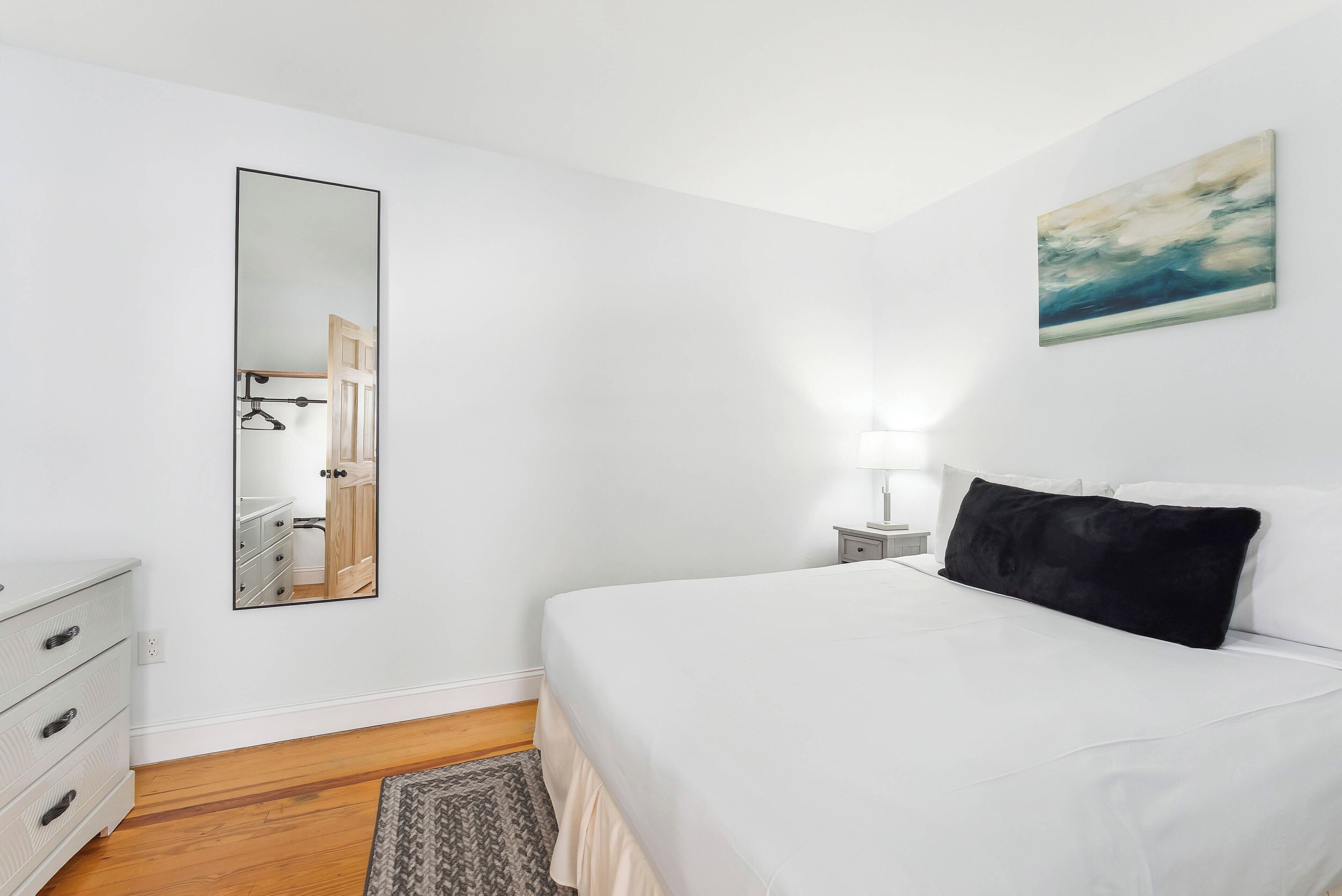 A bedroom with a bed, a dresser, a mirror, a nightstand, and a painting on the wall.