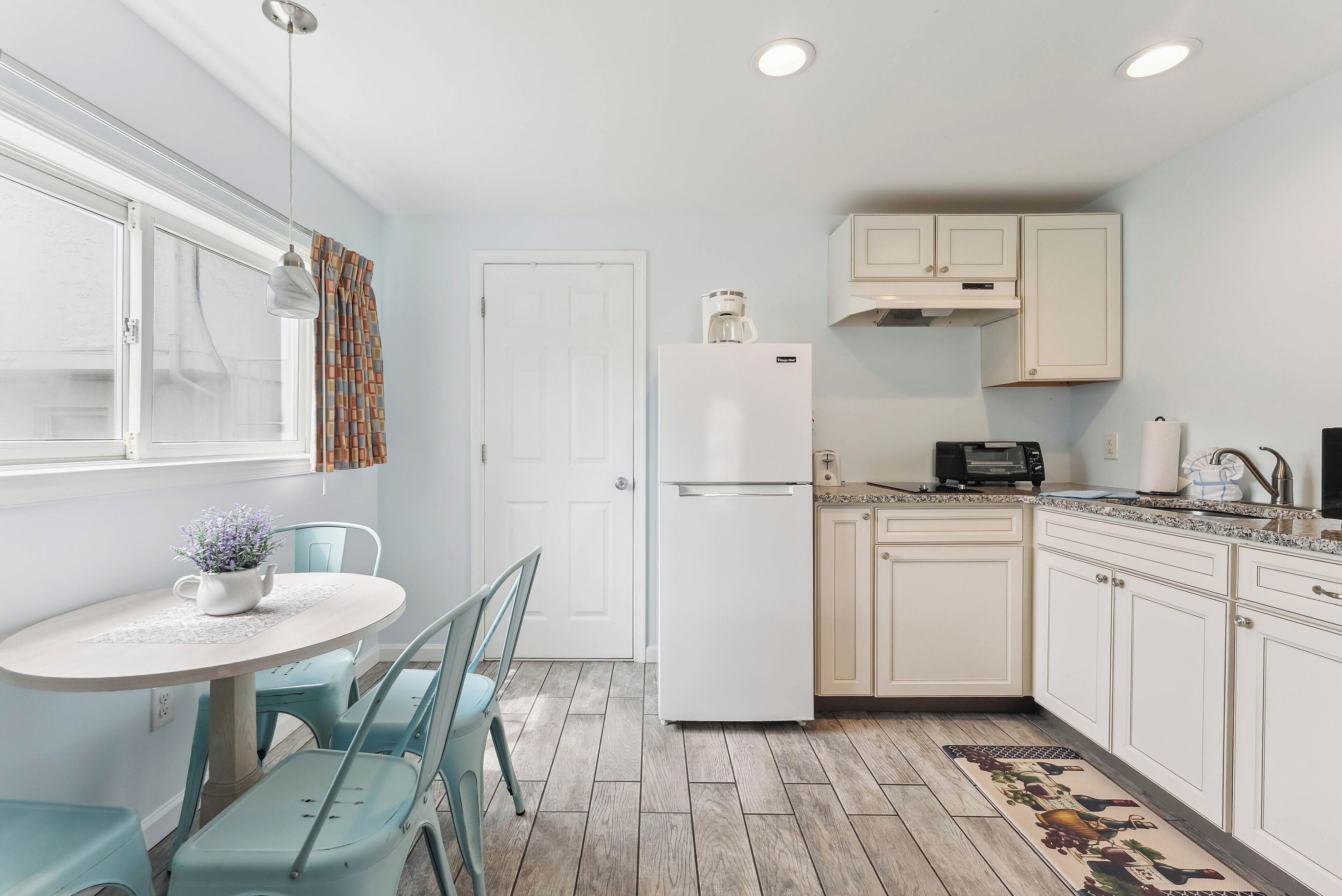 Cocina compacta con armarios blancos, un refrigerador, una mesa de comedor con sillas y un ventanal con cortinas.