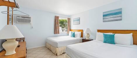 A hotel room with two beds, a wooden dresser, a lamp, an air conditioning unit, and a window with curtains.