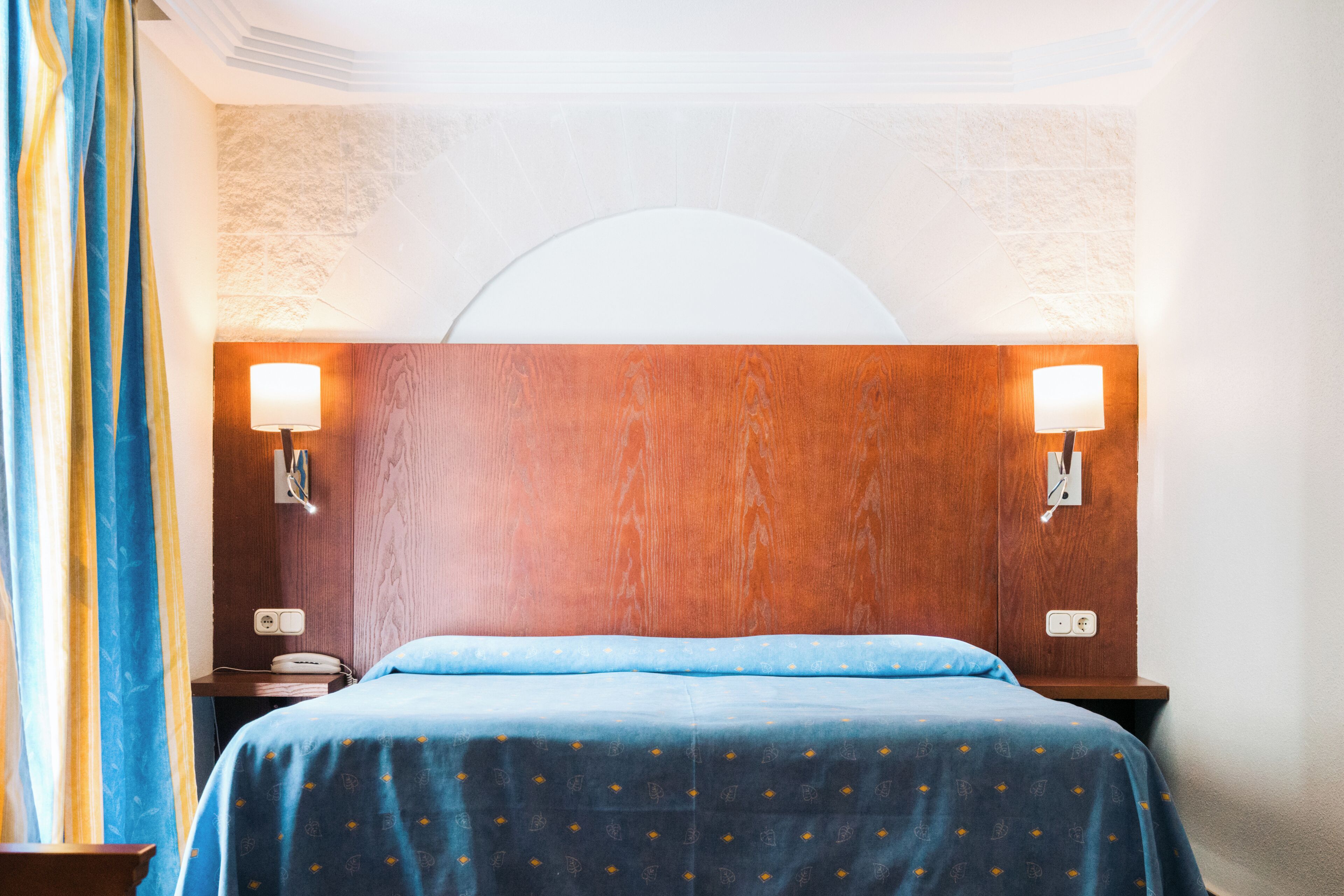 A hotel room with a wooden headboard, two bedside lamps, a bed with a blue bedspread, and a nightstand with a phone.