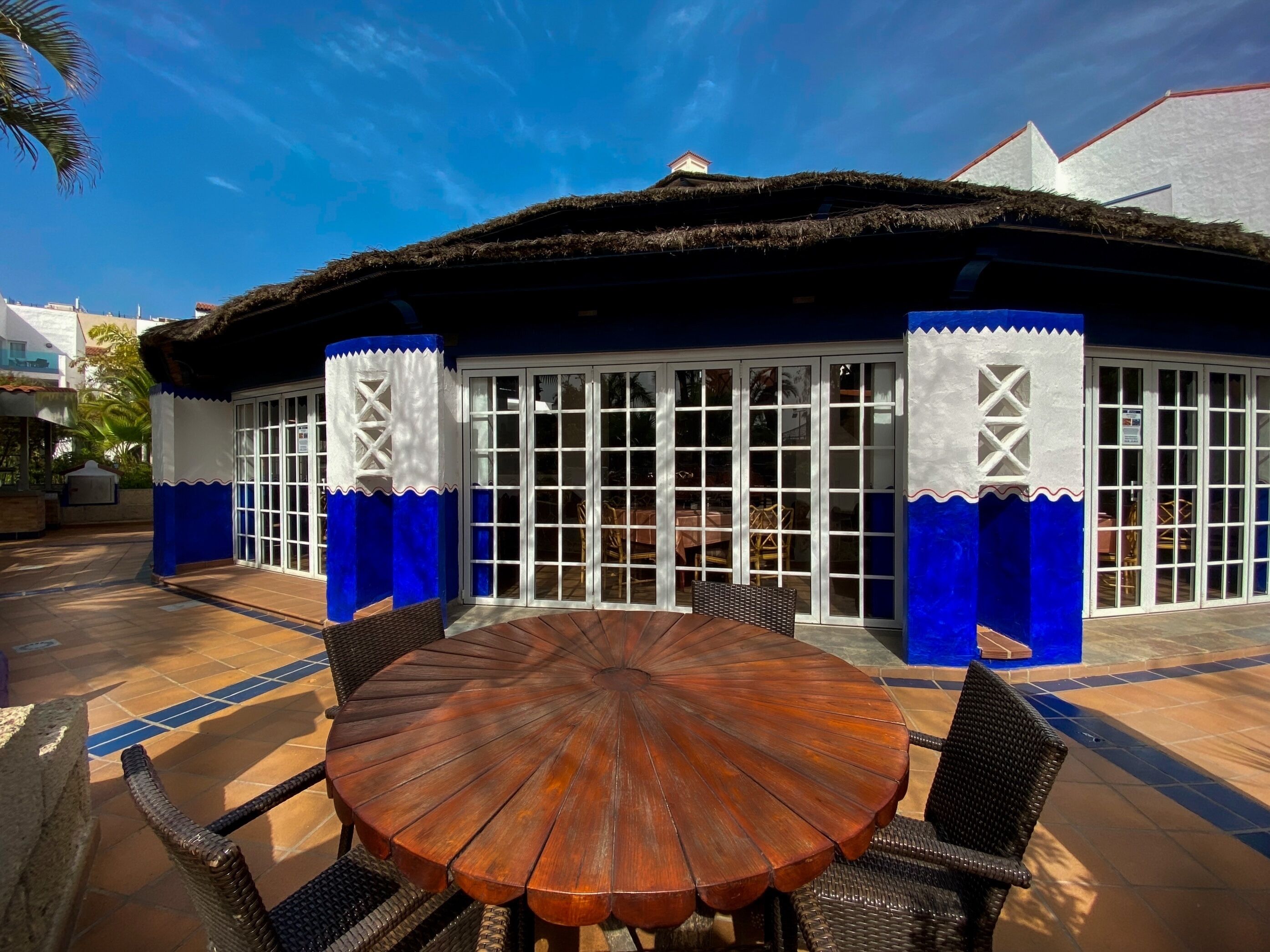 A covered outdoor seating area with a round wooden table and wicker chairs, a thatched roof, and a blue and white exterior.