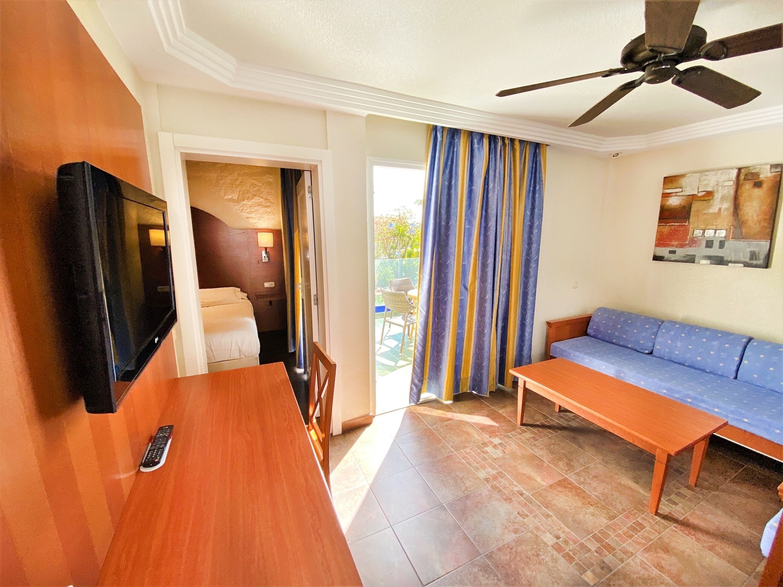 A hotel room with a bed, a TV, a ceiling fan, a sofa, and a table.