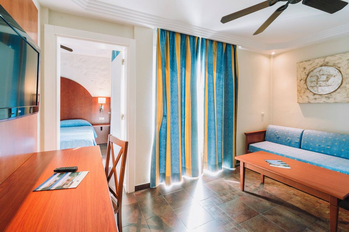 Une chambre d’hôtel avec une table en bois, un canapé, une télévision et un lit doté d’une tête de lit bleue.