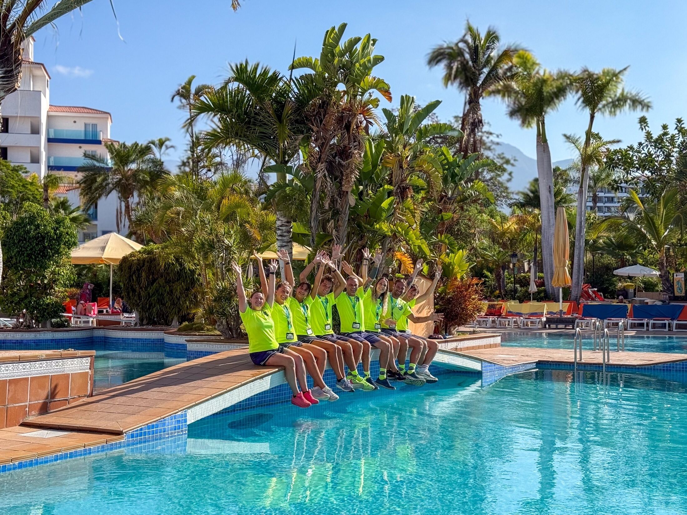 A group of people sitting on the edge of a pool with palm trees and a tiled deck in the background.