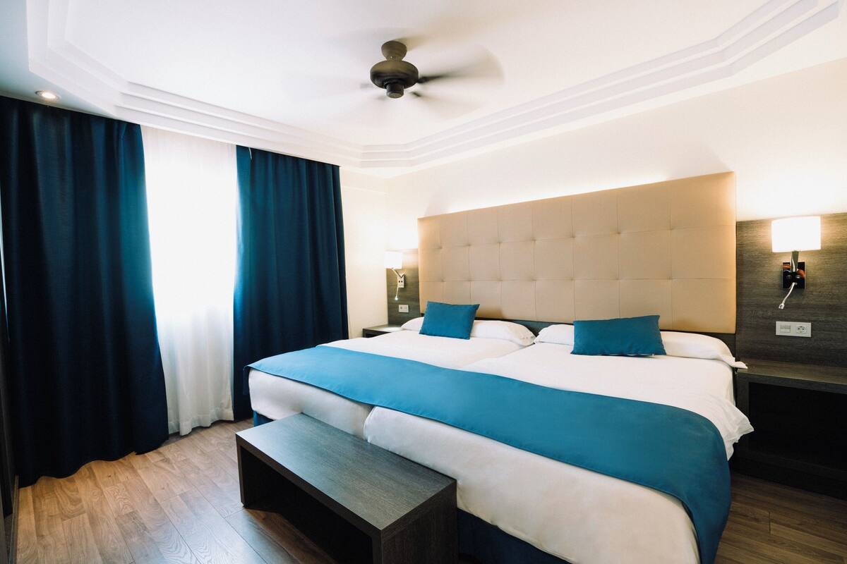 Une chambre d’hôtel avec un grand lit, une table de chevet, une commode, un ventilateur de plafond et une fenêtre avec des rideaux.