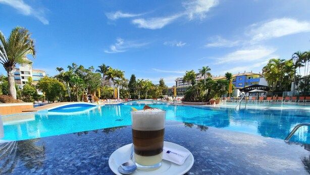 A clear view of a swimming pool with lounge chairs and palm trees, a glass of a layered beverage on a table, and a sign reading "INFOCENTER". - Park Club Europe - All Inclusive (Arona)