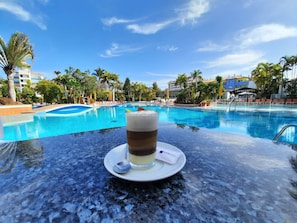 A clear view of a swimming pool with lounge chairs and palm trees, a glass of a layered beverage on a table, and a sign reading "INFOCENTER".