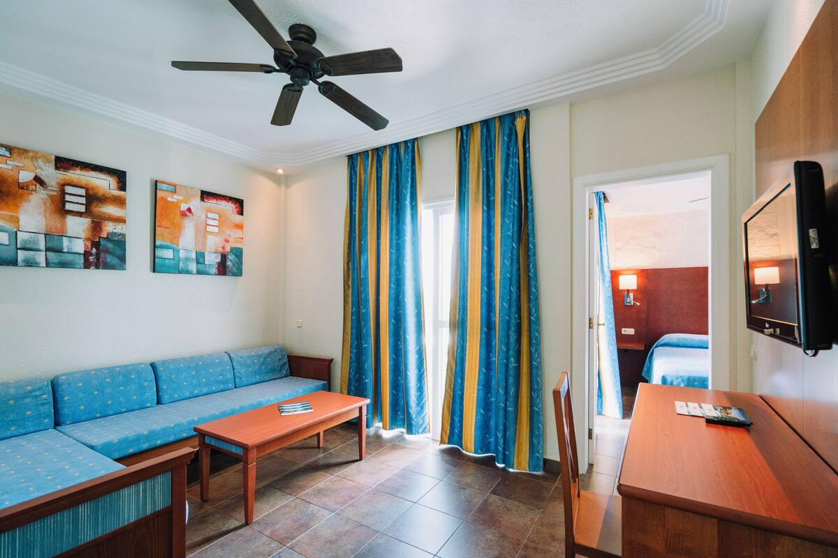 Une chambre d’hôtel avec un canapé, une table en bois, un ventilateur de plafond et des rideaux.