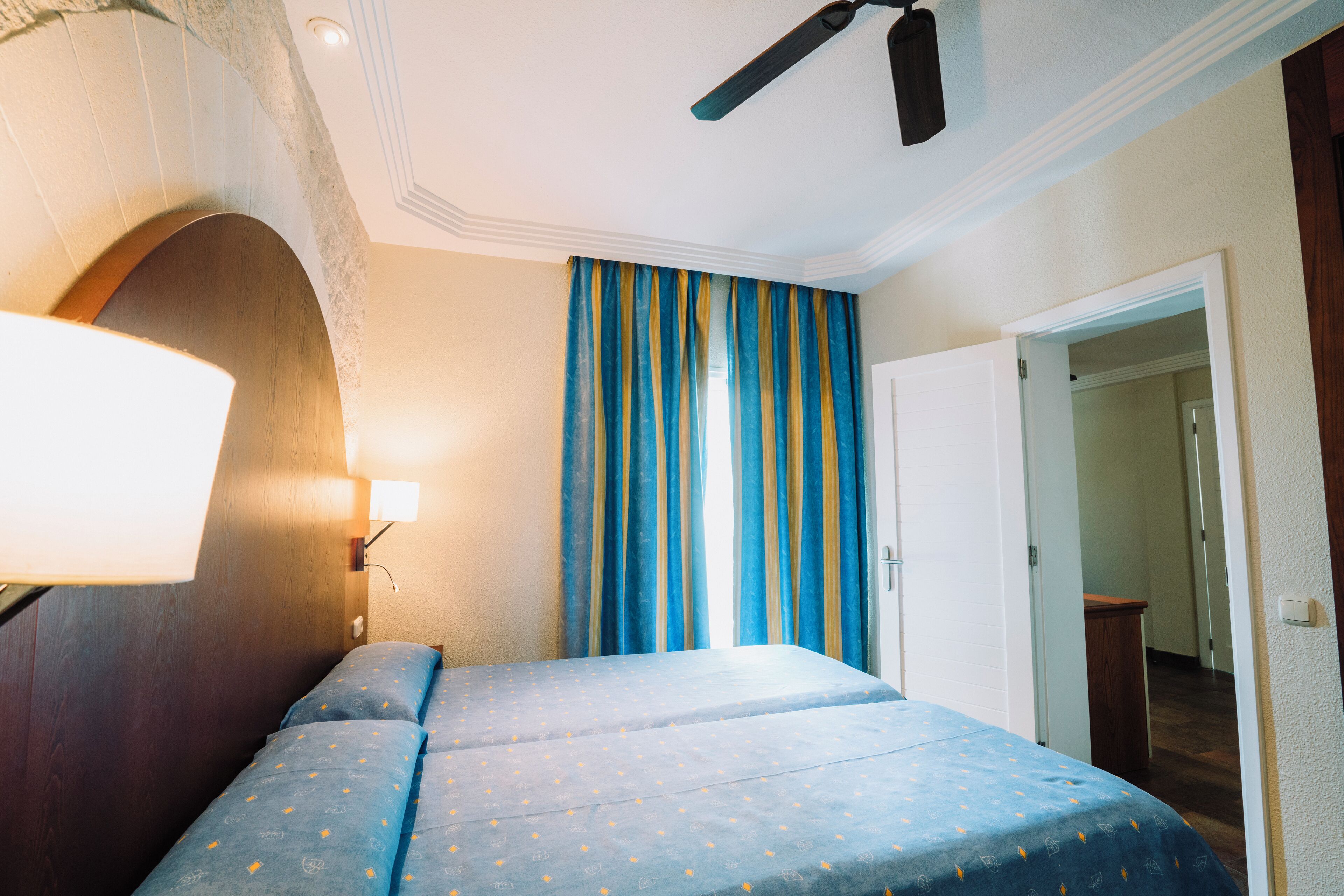 A bedroom with a bed, a ceiling fan, a lamp, and curtains.