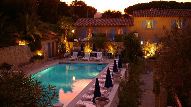 A pool area with lounge chairs and umbrellas, a stone wall, and a building with a tiled roof in the background. - Le Jas Neuf (Sainte-Maxime)
