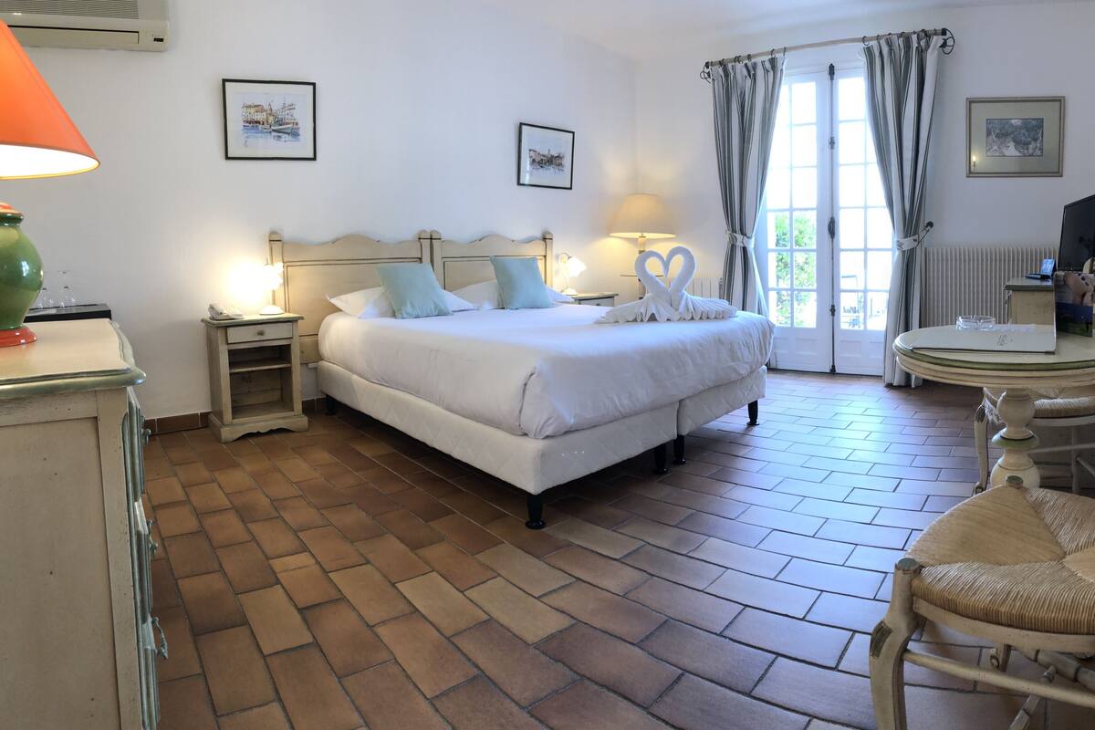 Une chambre d’hôtel avec un grand lit, deux lampes de chevet, une petite table, une chaise et une fenêtre avec des rideaux.