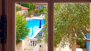 A view of a swimming pool with a blue umbrella, surrounded by greenery and a stone building. - Le Jas Neuf (Sainte-Maxime)
