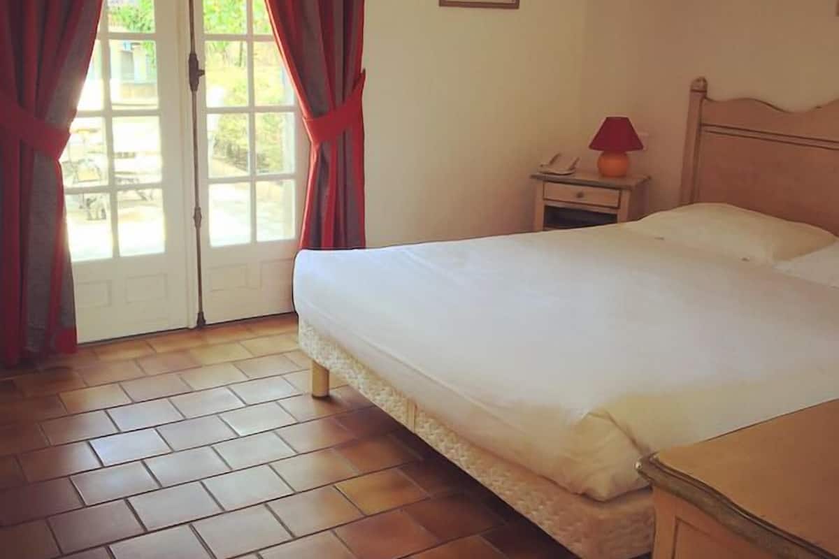 Une chambre à coucher avec un lit, une table de chevet, une lampe et une fenêtre avec des rideaux.