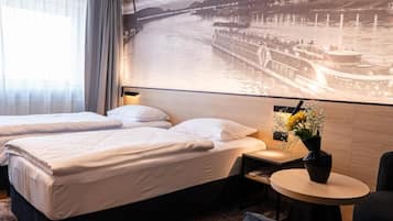 A hotel room with two beds, a small table with a vase of flowers, and a large wall mural of a historic building by a river.