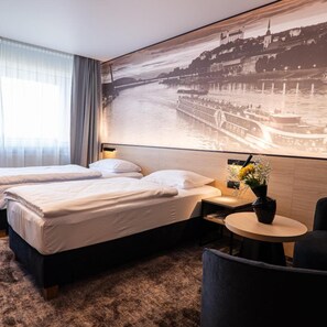 Une chambre d’hôtel avec deux lits, une petite table ornée d’un vase de fleurs et une grande fresque murale représentant un bâtiment historique situé au bord d’une rivière.