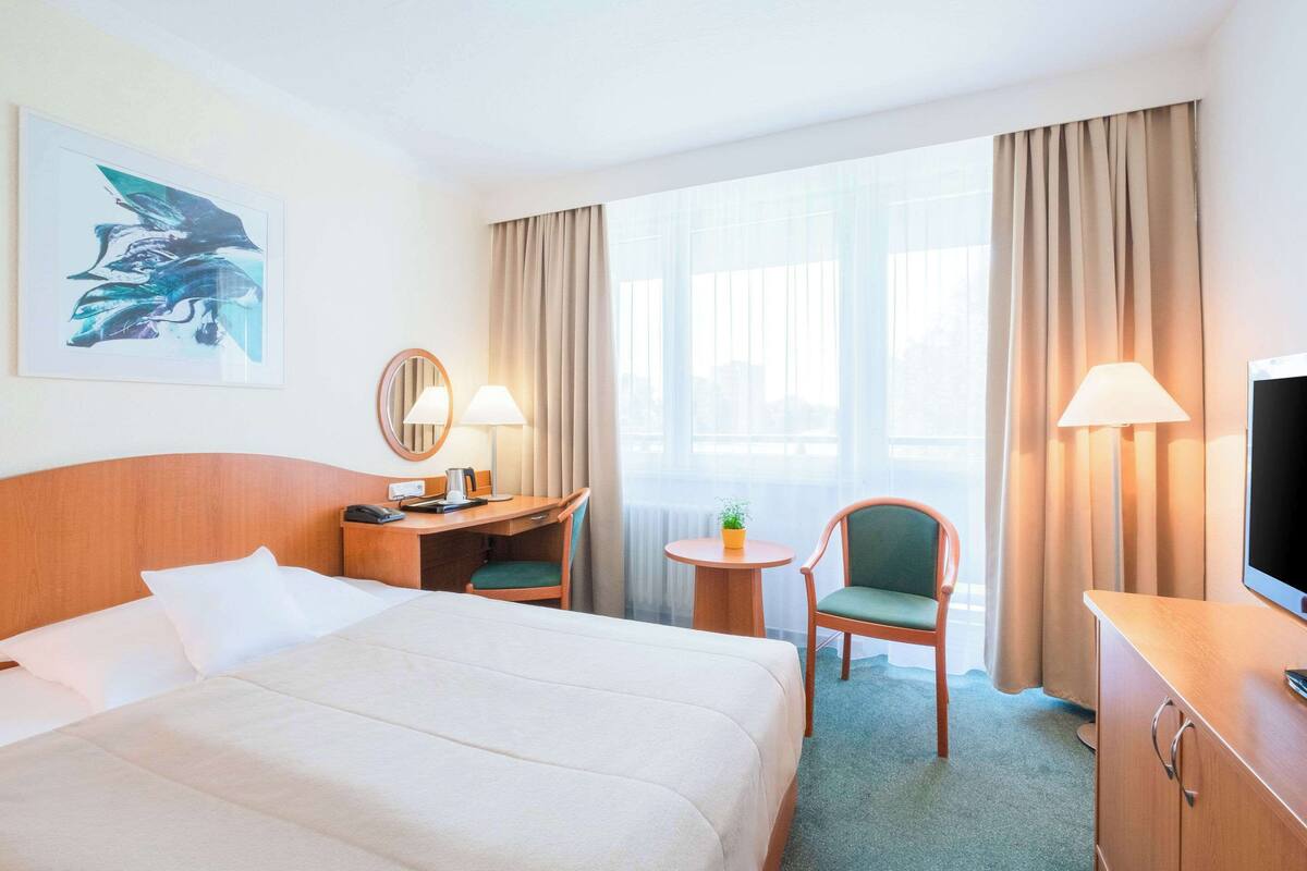 Ein Hotelzimmer mit einem großen Bett, einem Schreibtisch mit Lampe, einem Sessel, einem kleinen Tisch mit Pflanze und einem Fernseher.