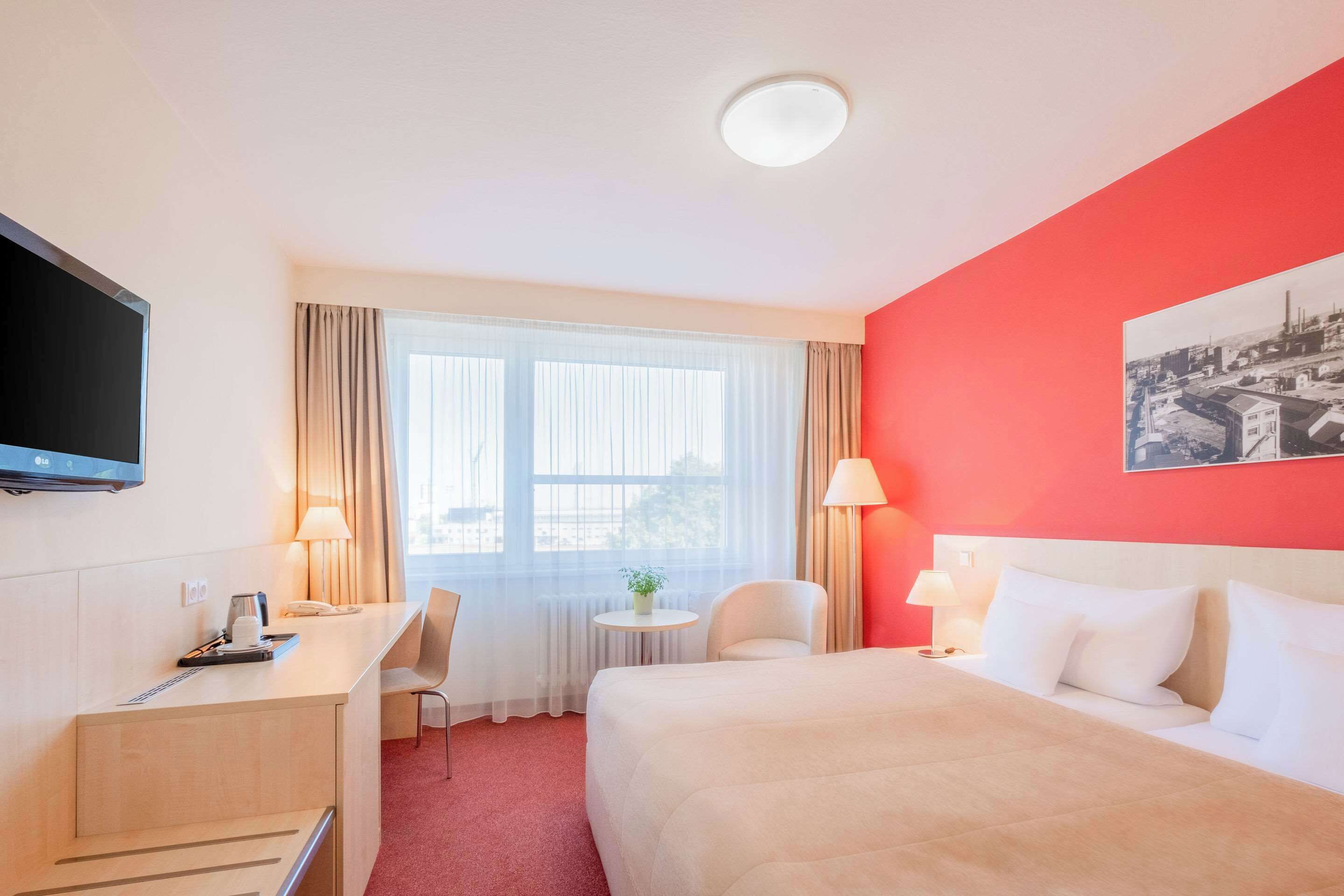 Quarto de hotel com cama, escrivaninha, cadeira, televisão e janela com cortinas.