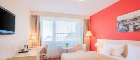 Quarto de hotel com cama, escrivaninha, cadeira, televisão e janela com cortinas.