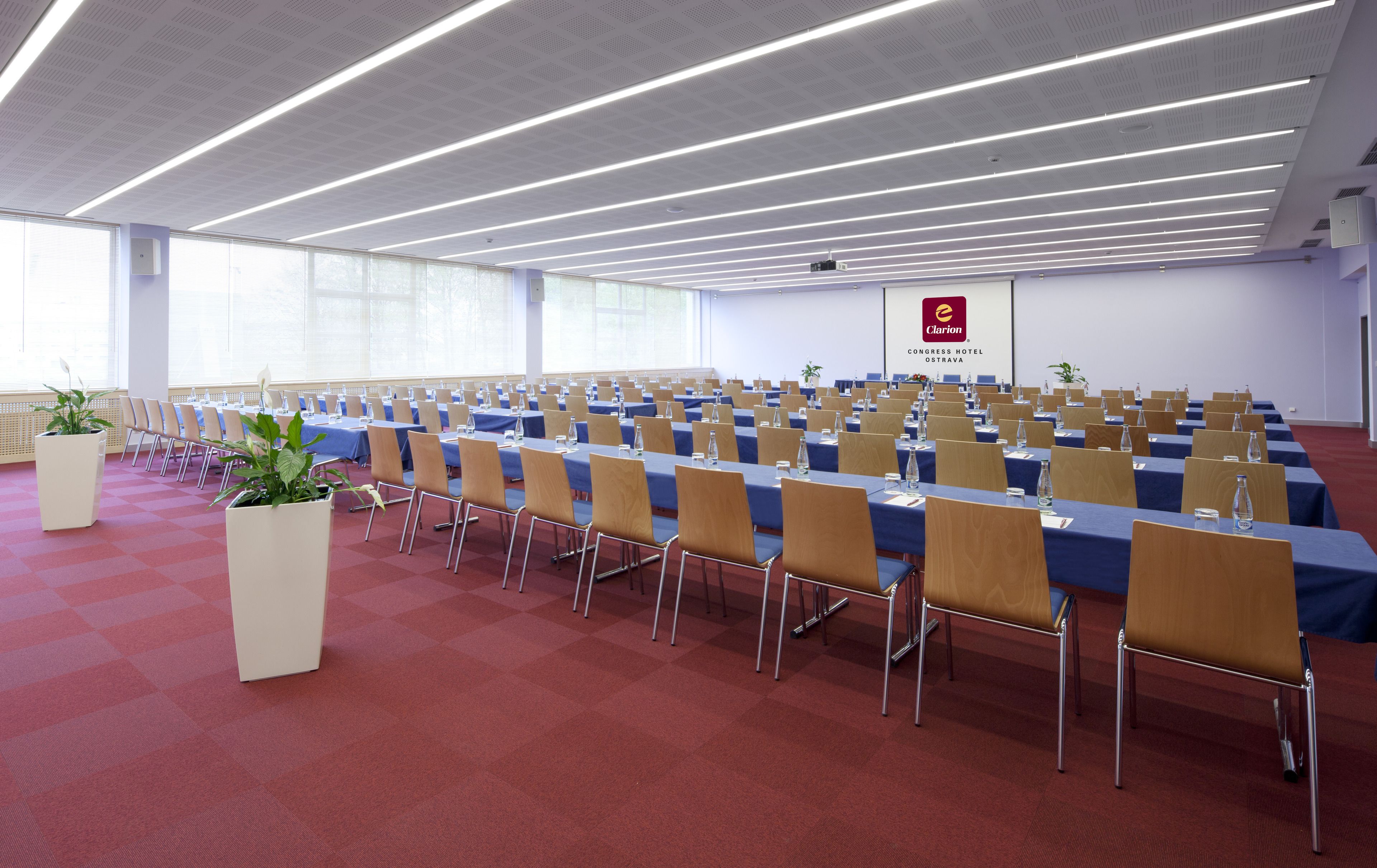 Uma sala de conferência com fileiras de mesas e cadeiras, janelas amplas e luminárias lineares no teto.