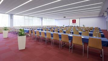 Uma sala de conferência com fileiras de mesas e cadeiras, janelas amplas e luminárias lineares no teto.