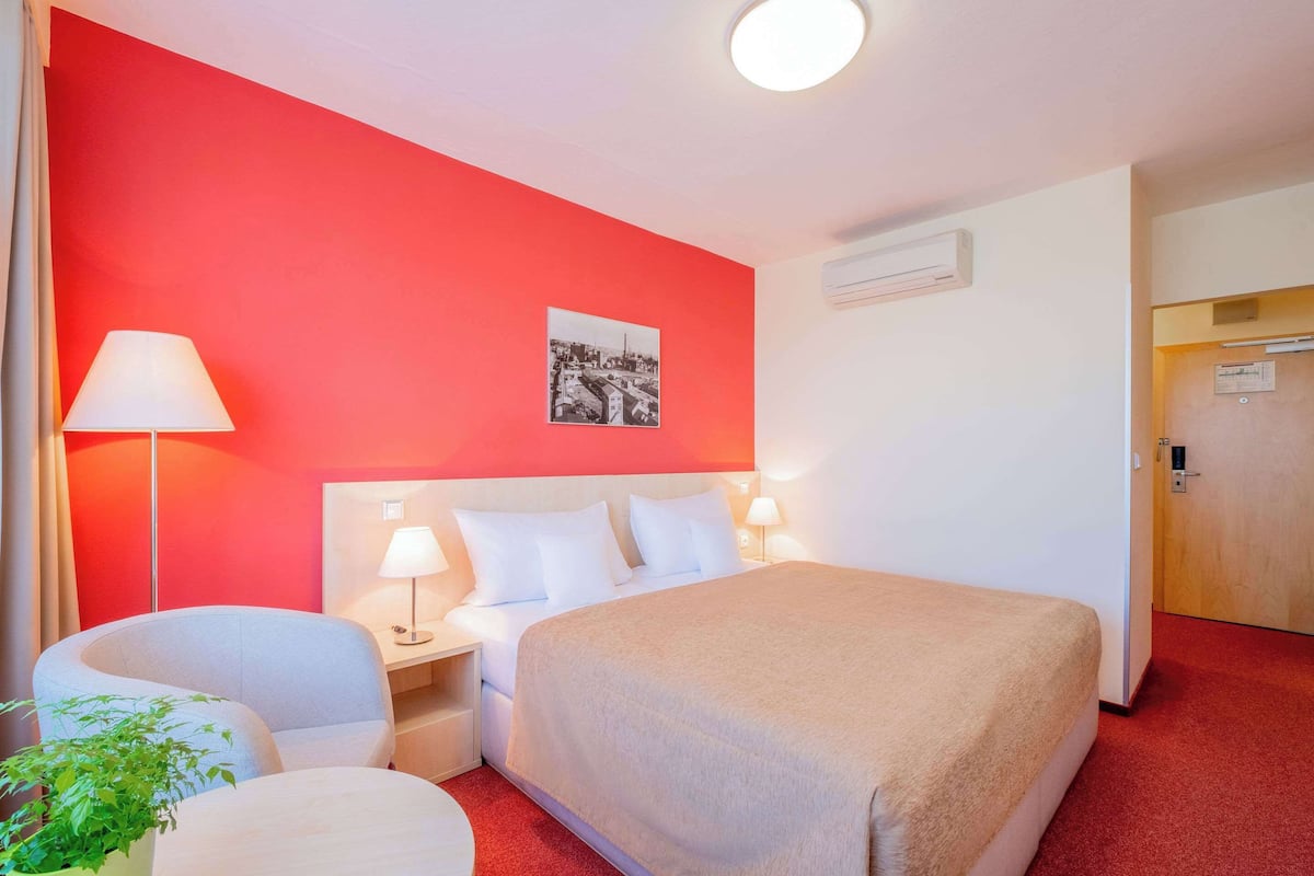 Ein Hotelzimmer mit roter Akzentwand, einem Bett mit weißer Bettwäsche, einem Schreibtisch mit Lampe und einem kleinen runden Tisch mit einer Pflanze.