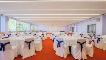 Un salón amplio con filas de mesas y sillas blancas, cada una con un lazo azul, y un suelo cubierto de alfombra roja.