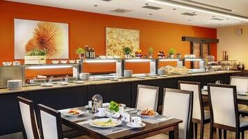 Espaço de pequeno-almoço estilo buffet, com um balcão comprido, várias estações de serviço e mesas com cadeiras.