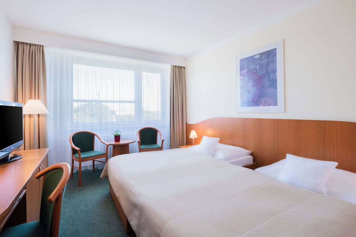 Ein Hotelzimmer mit einem großen Bett, zwei Sesseln, einem Schreibtisch mit Fernseher, einem kleinen Tisch und einem Fenster mit Vorhängen.