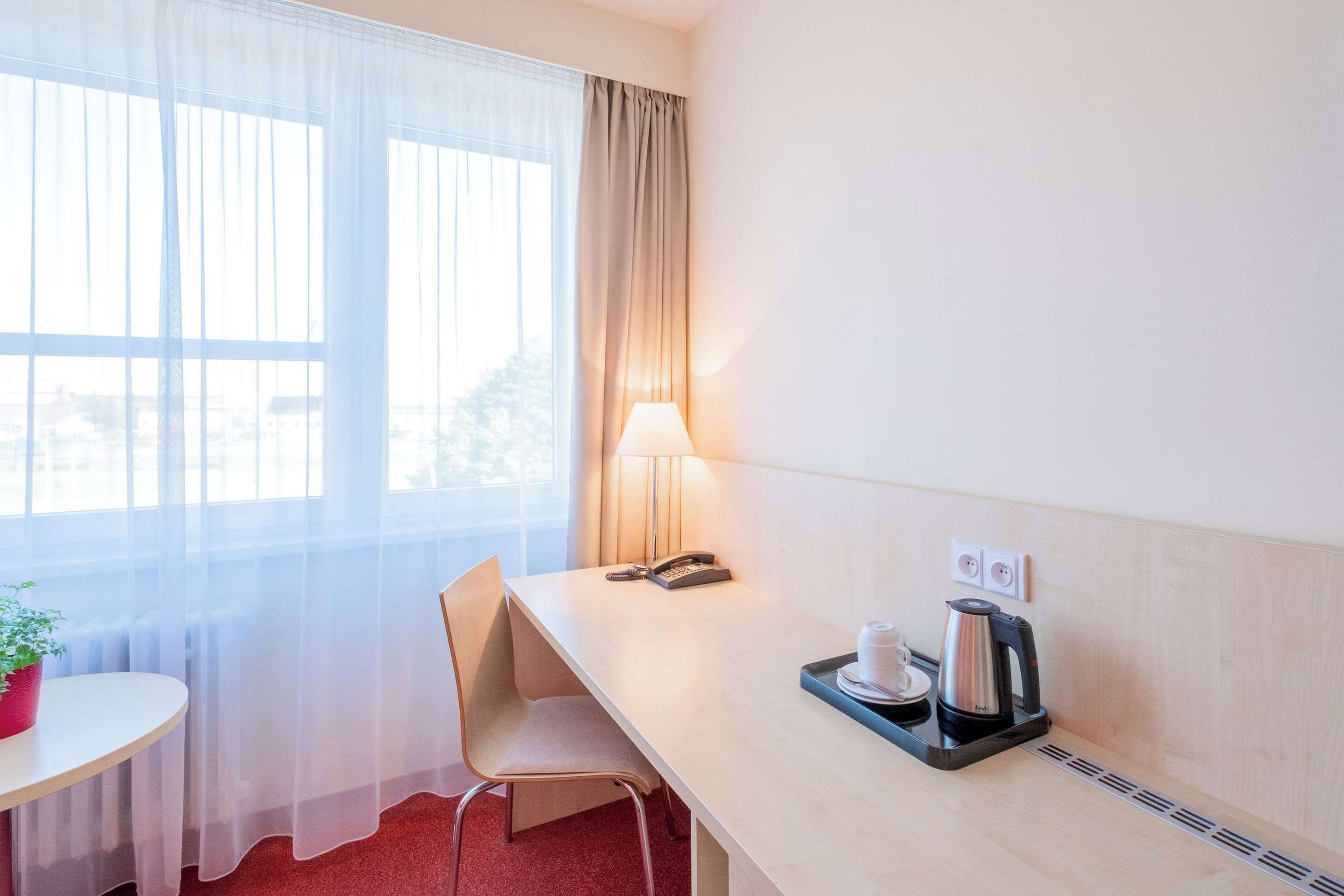 Ein Hotelzimmer mit Schreibtisch, Stuhl, Lampe, Telefon, Kaffeemaschine und einem kleinen Tisch mit Topfpflanze.