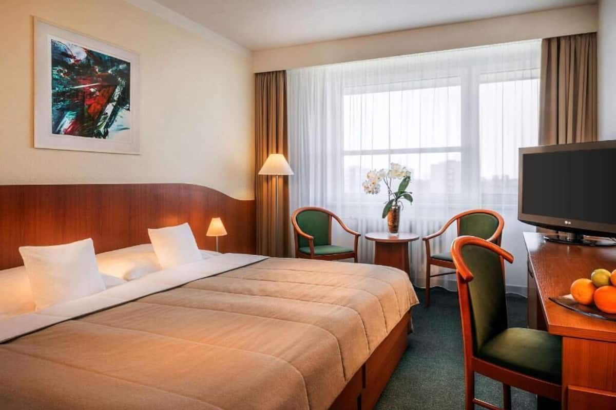 Ein Hotelzimmer mit einem Bett, einem Schreibtisch mit Fernseher, einem Sessel, einem kleinen Tisch mit Blumen und einem Fenster mit Vorhängen.