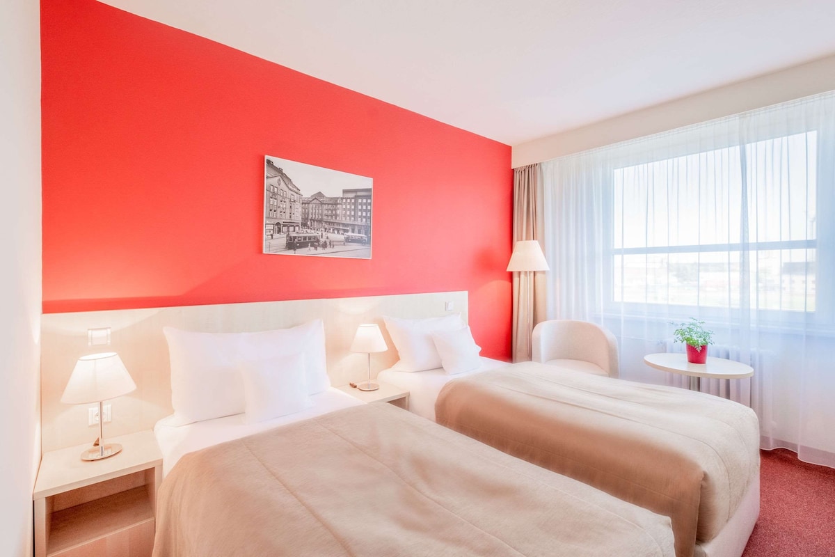 A hotel room with two beds, a red accent wall, a window with sheer curtains, a small round table with a plant, and a wall-mounted picture.