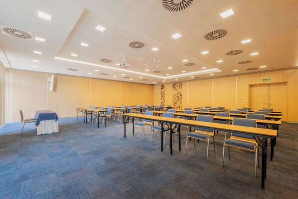 Un salón de conferencias con mesas y sillas dispuestas en filas, luces en el techo y una puerta.