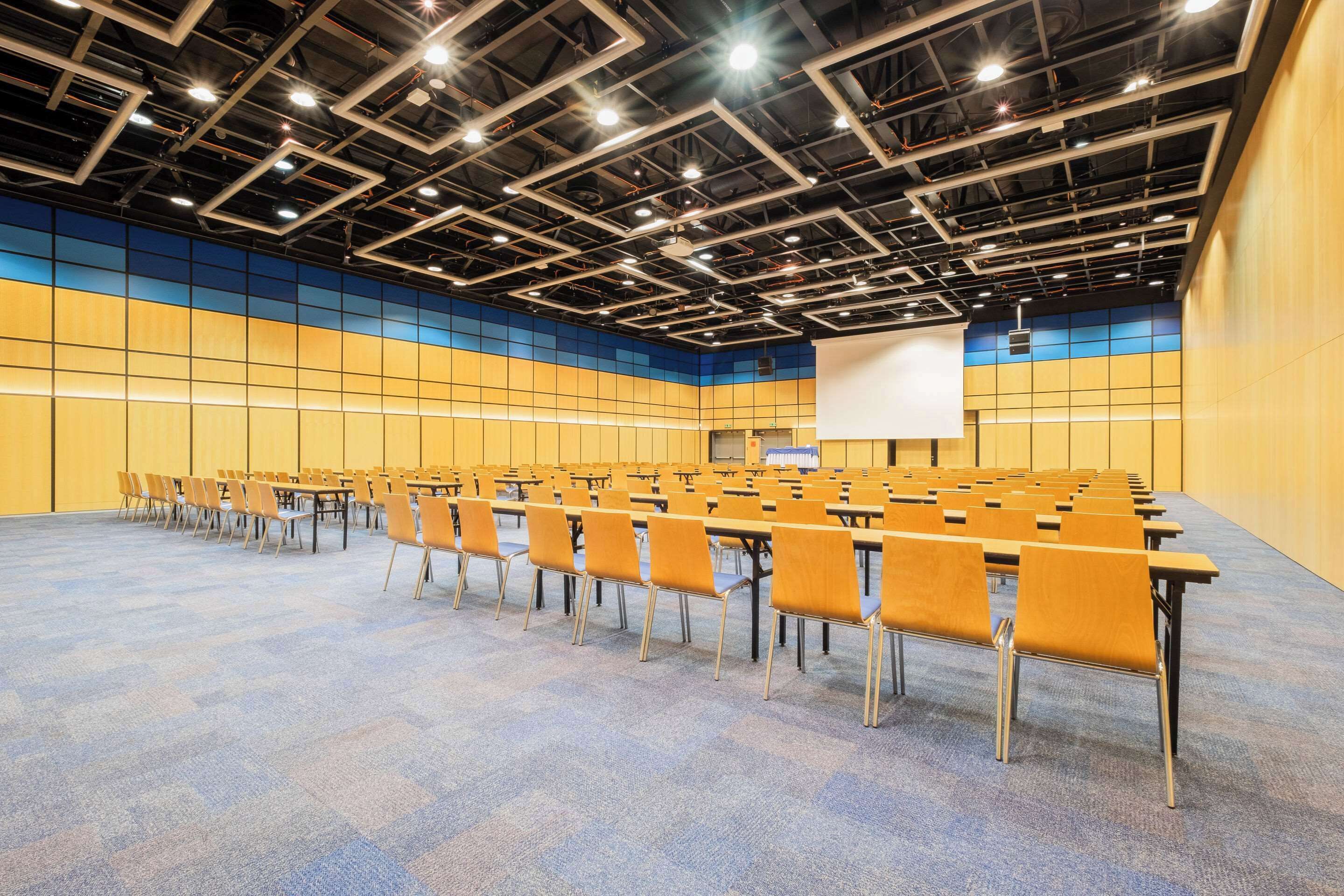 A conference room with rows of chairs and tables, a projector screen, and a ceiling with exposed pipes and lighting fixtures.
