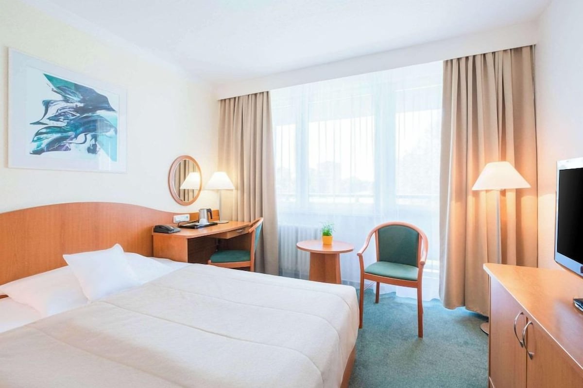 A hotel room with a bed, desk, chair, TV, and large windows.