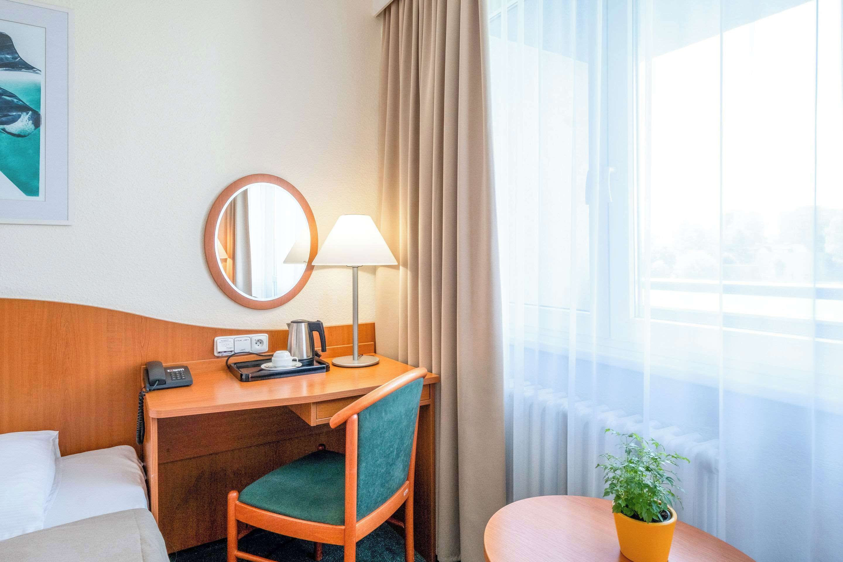 A hotel room with a bed, a desk with a lamp, a telephone, a coffee maker, and a potted plant.