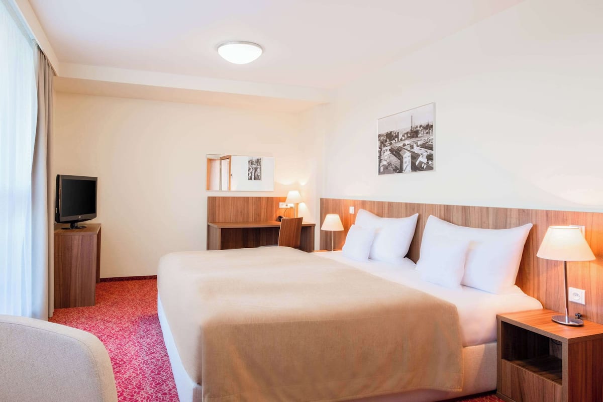 Ein Hotelzimmer mit einem großen Bett, einem Schreibtisch mit Computer, einem Fernseher und einem Bild an der Wand.