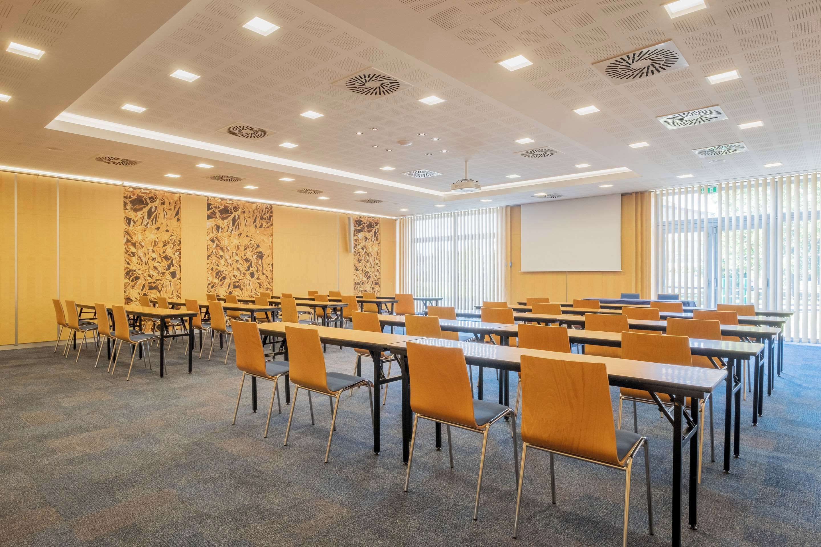 A conference room with tables and chairs arranged for meetings, a projector screen, and a large window with blinds.