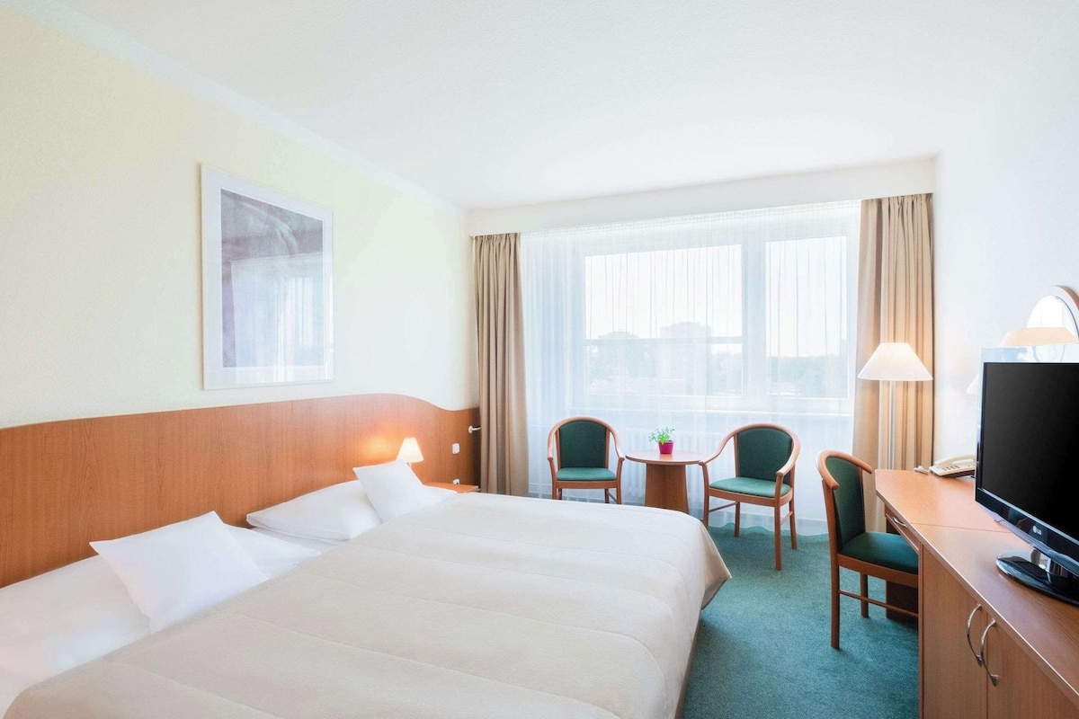 A hotel room with a large bed, two chairs, a desk with a TV, and a window with curtains.
