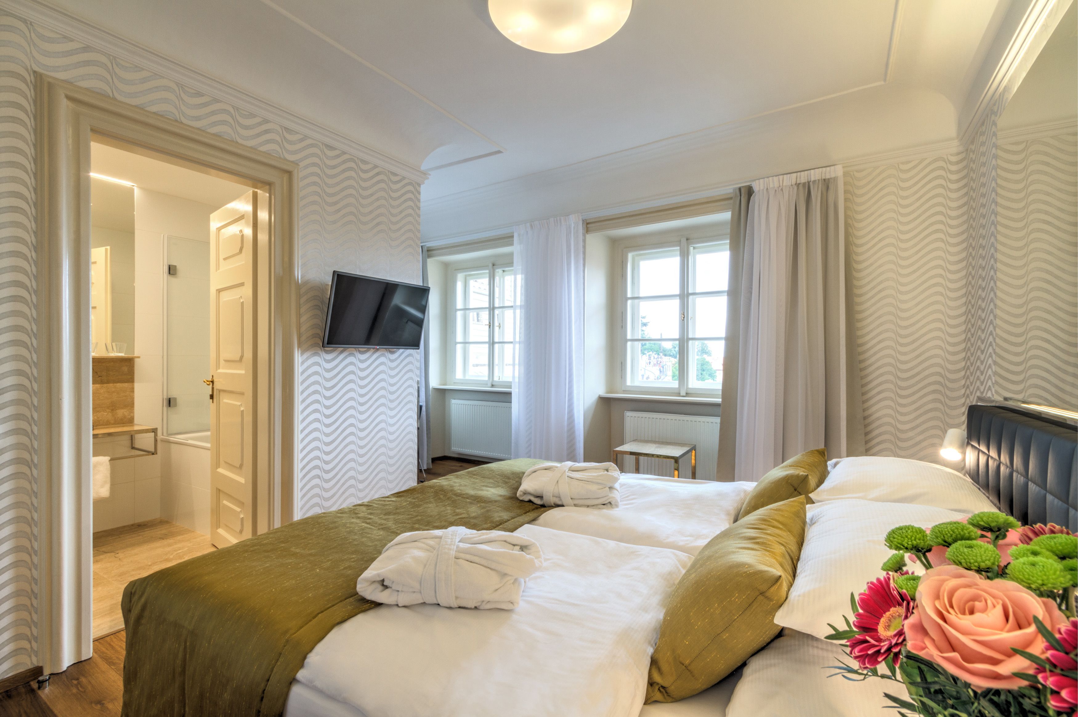 A hotel room with a bed, a TV, a window with curtains, and a bathroom visible through an open door.