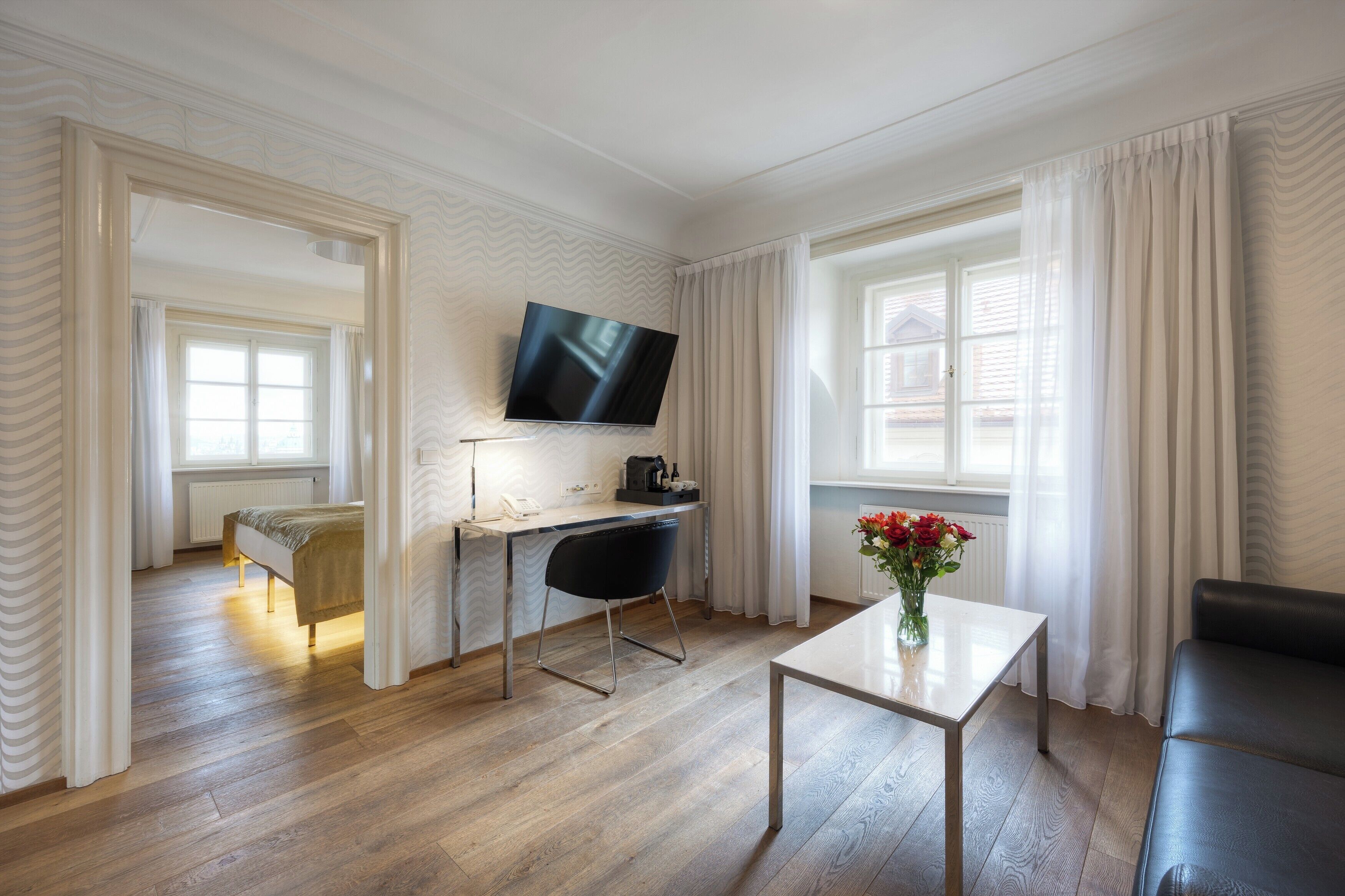 Moderni hotellihuone, jossa on puulattia, sänky, työpöytä ja taso-tv, sohva sekä maljakko, jossa on kukkia.