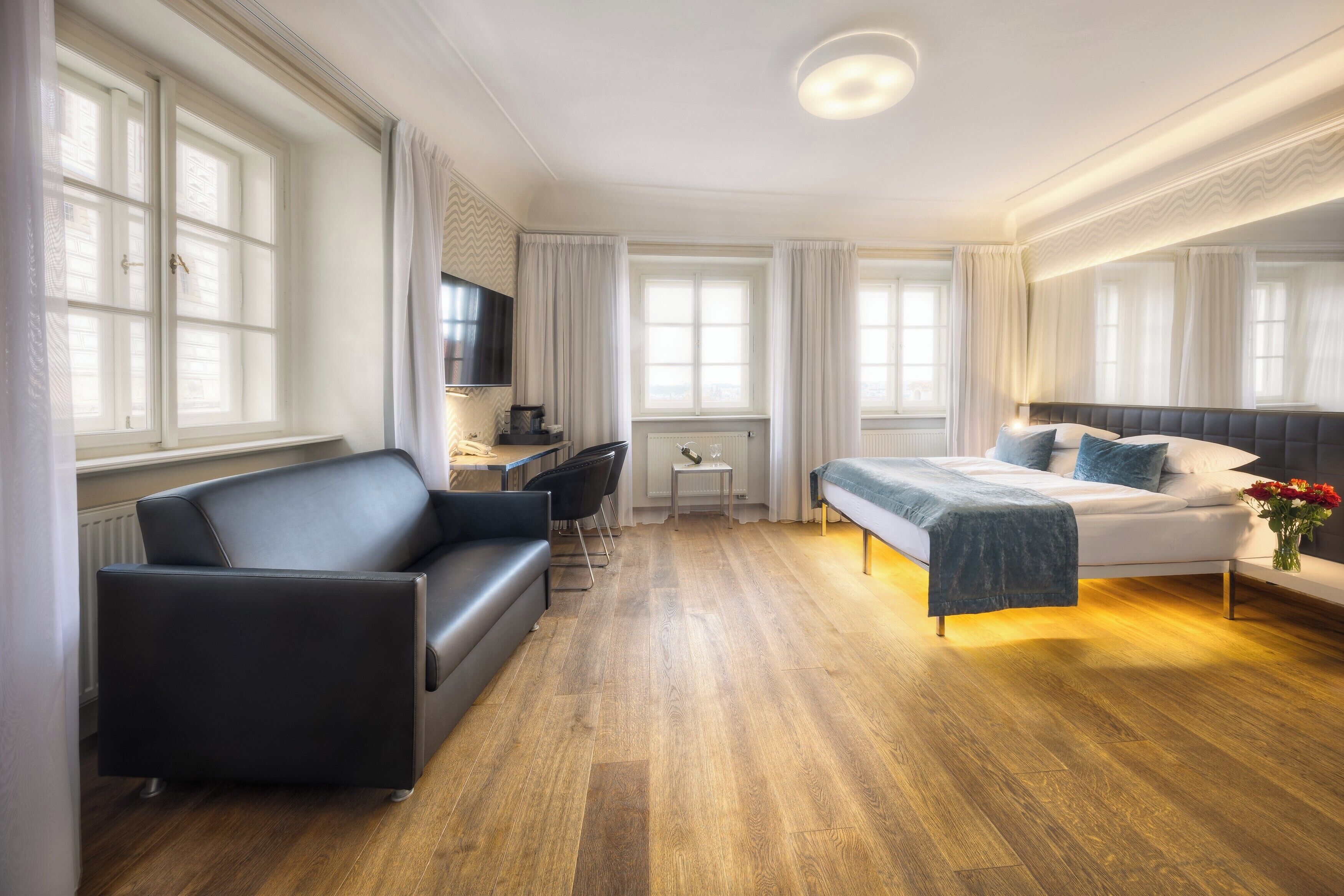 A modern hotel room with a bed, a sofa, a desk, and large windows.