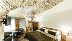 A room with a rustic stone ceiling, a bed with a wooden headboard, a small table with a lamp, and a television mounted on the wall. - Archibald At the Charles Bridge (Prague)