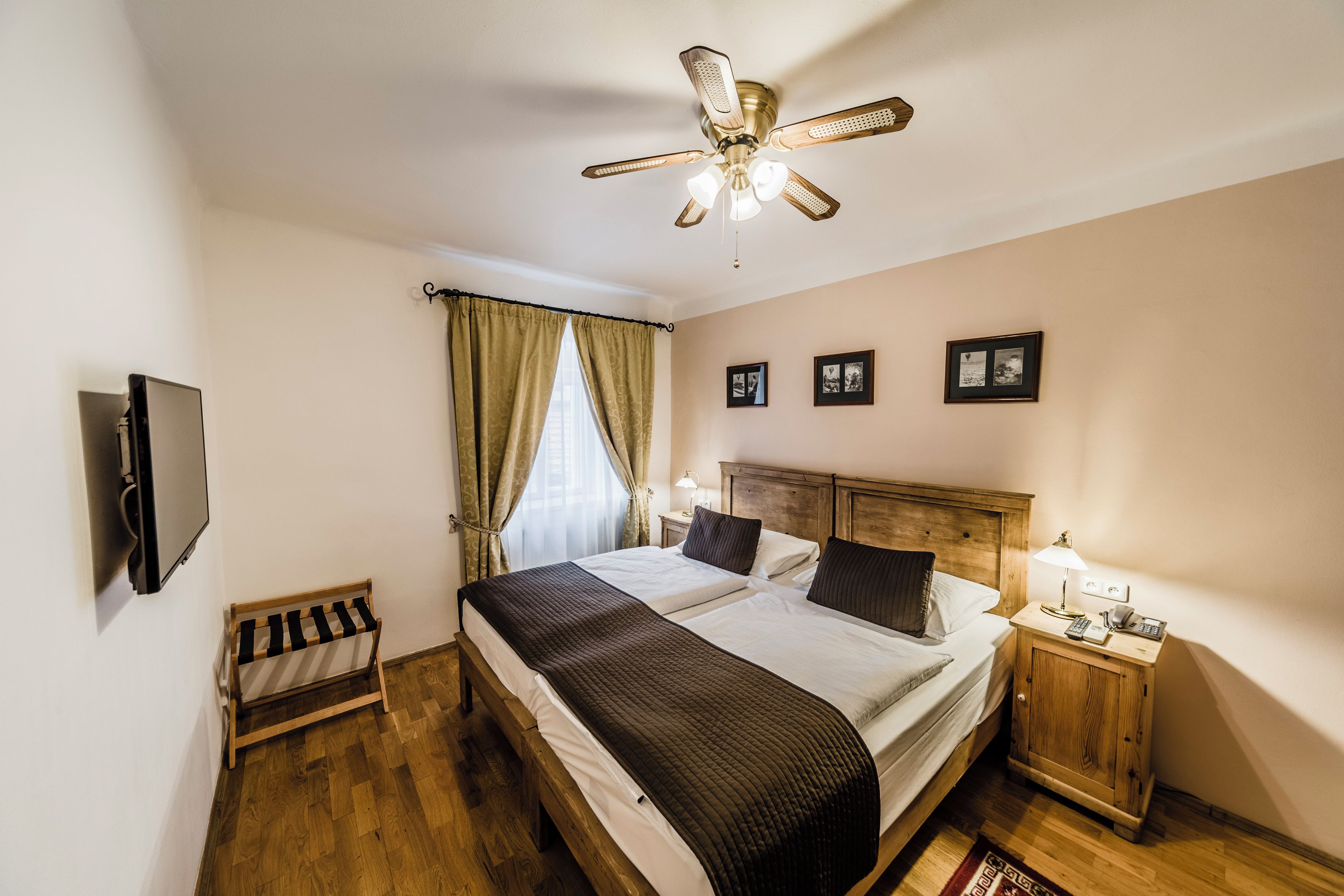 Un dormitorio con cama, mesita de noche, televisor, ventilador de techo y ventana con cortinas.