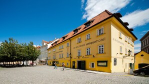 A yellow building with "At the Charles Bridge" and "an archibald uniquehotel" signage, a cobblestone street, and a clear blue sky. - Archibald At the Charles Bridge (Prague)