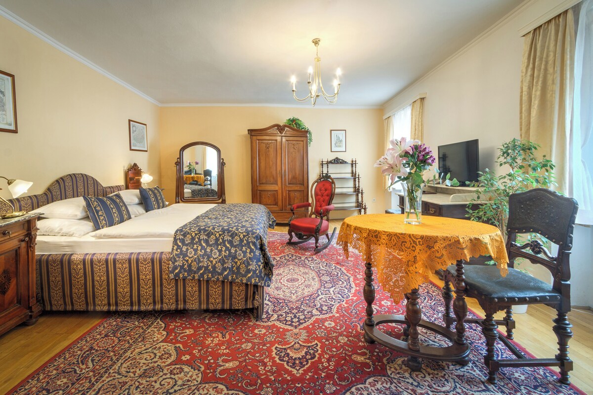 Een klassieke kamer met een bed, een houten garderobe, een schommelstoel, een tafel met een vaas en een televisie.